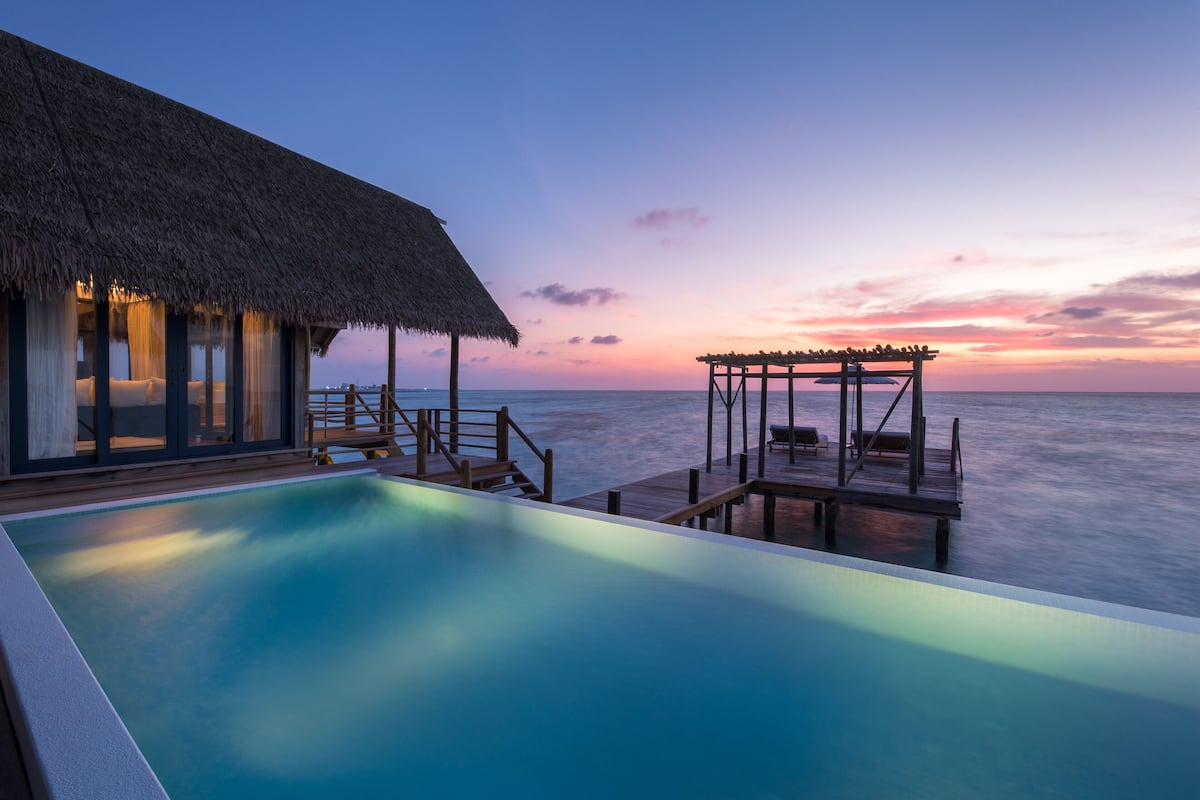 Ein Infinity-Pool mit Blick auf den Ozean bei Sonnenuntergang, ein strohgedecktes Gebäude und Sitzgelegenheiten auf einer Holzterrasse.
