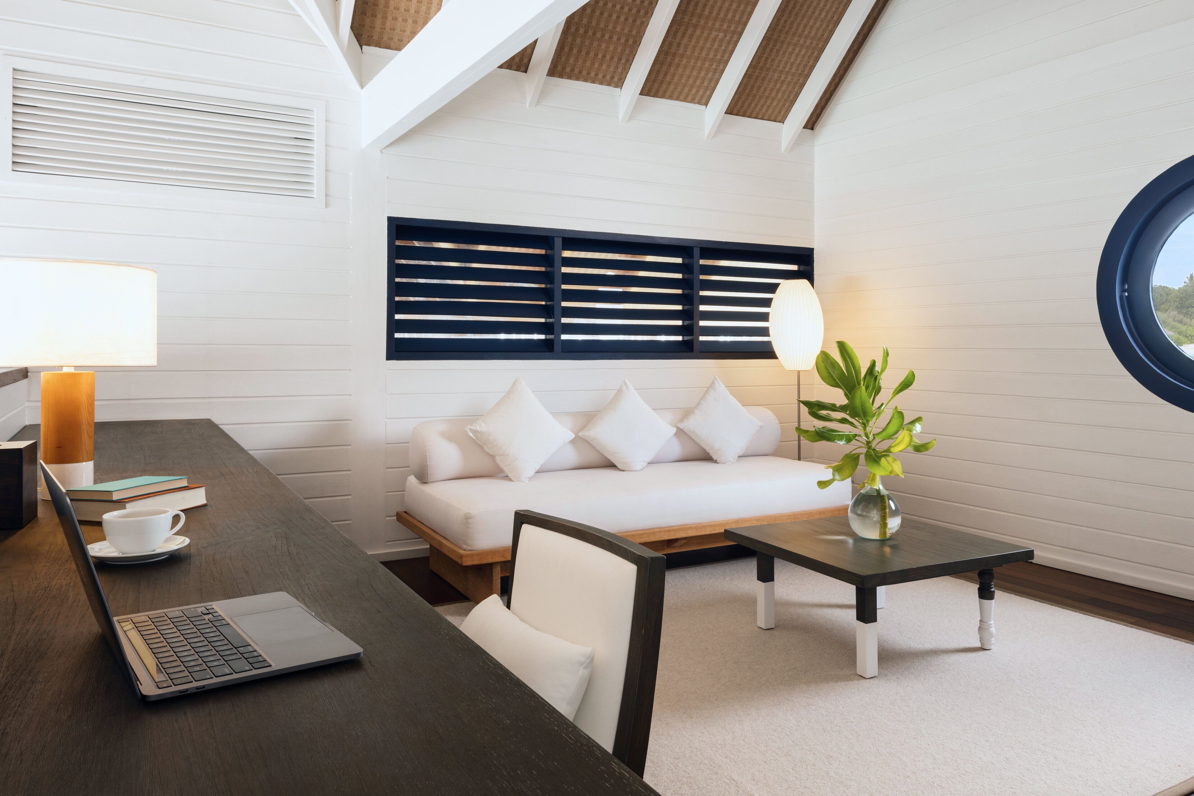 A modern room with a wooden desk, a laptop, a white sofa, a round window, and a plant on a small table.