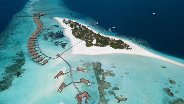 An aerial view of a resort with overwater bungalows connected by walkways, a sandy beach, and clear turquoise waters.