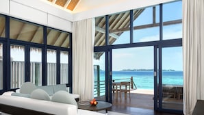 A modern room with a large window overlooking a beach, a sofa, a table with fruit, and a view of the ocean.