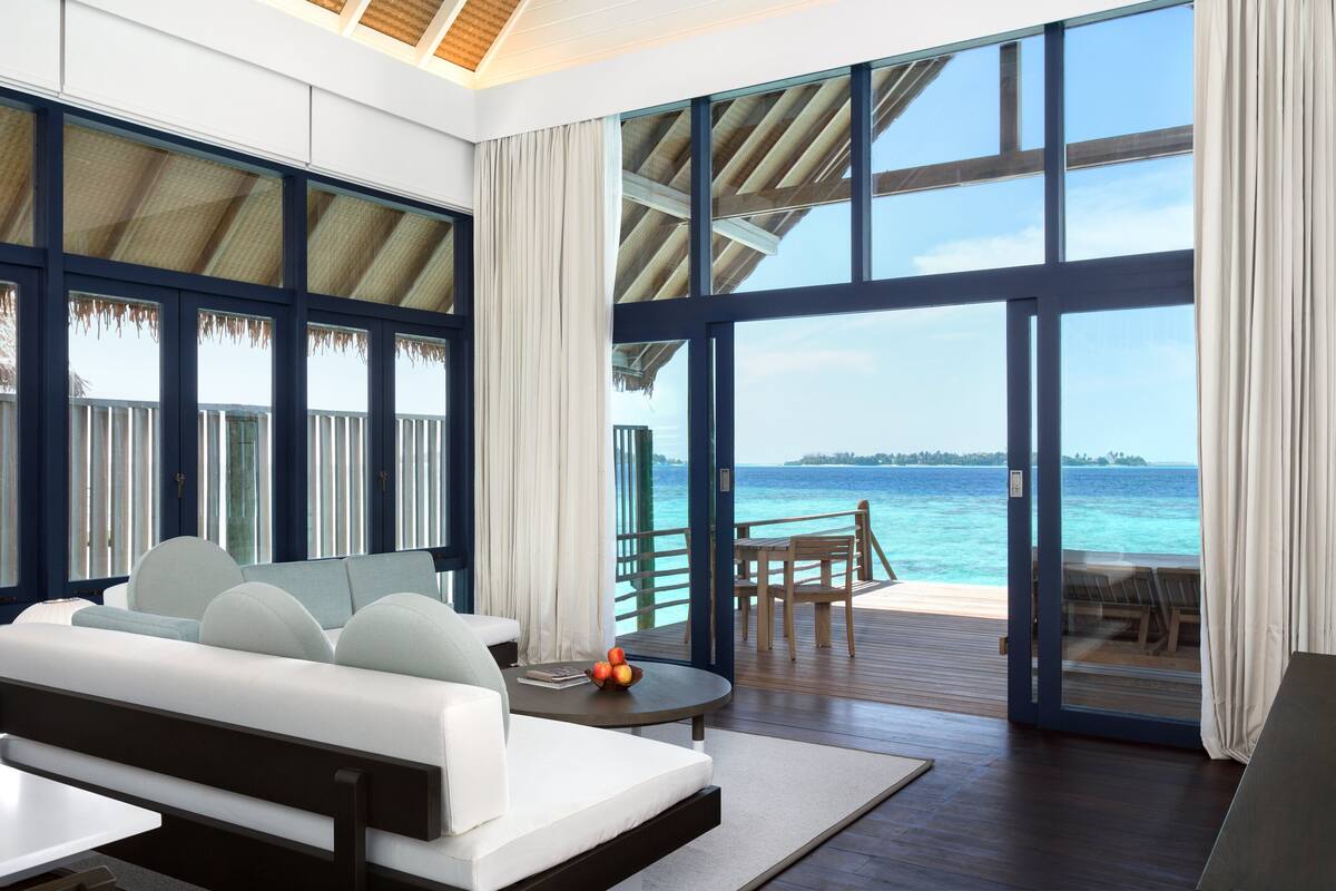 Ein modernes Zimmer mit großem Fenster mit Blick auf einen Strand, einer Couch, einem Tisch mit Obst und Meerblick.