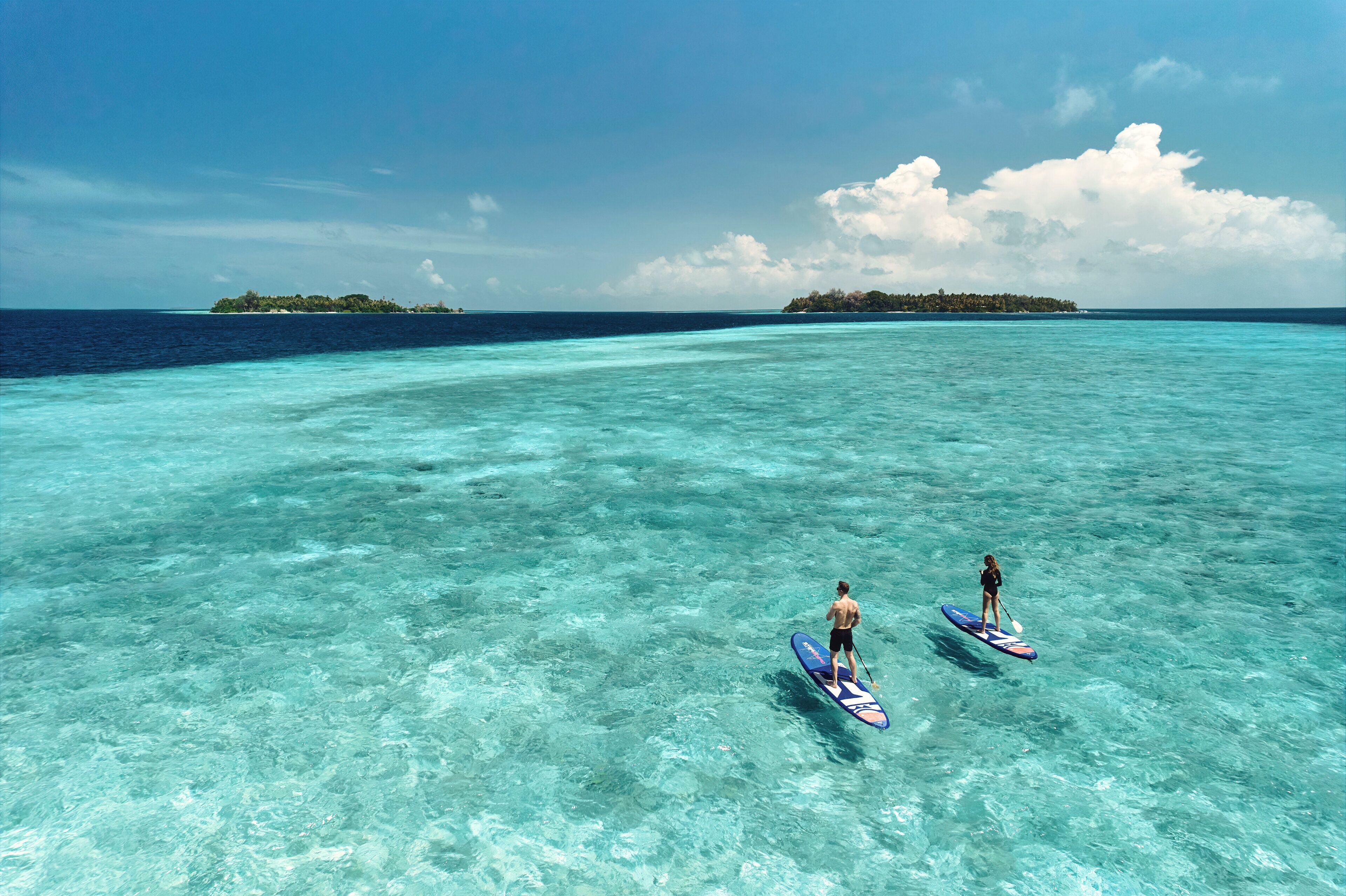Twee mensen staan op paddleboards op helder turquoise water, met een eiland in de verte.
