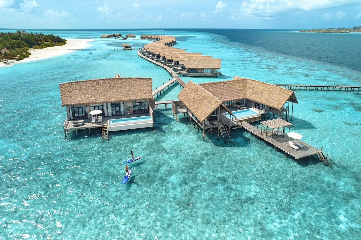Bungalows über dem Wasser mit Strohdächern, klares türkisblaues Wasser und ein Holzsteg, der sie verbindet.