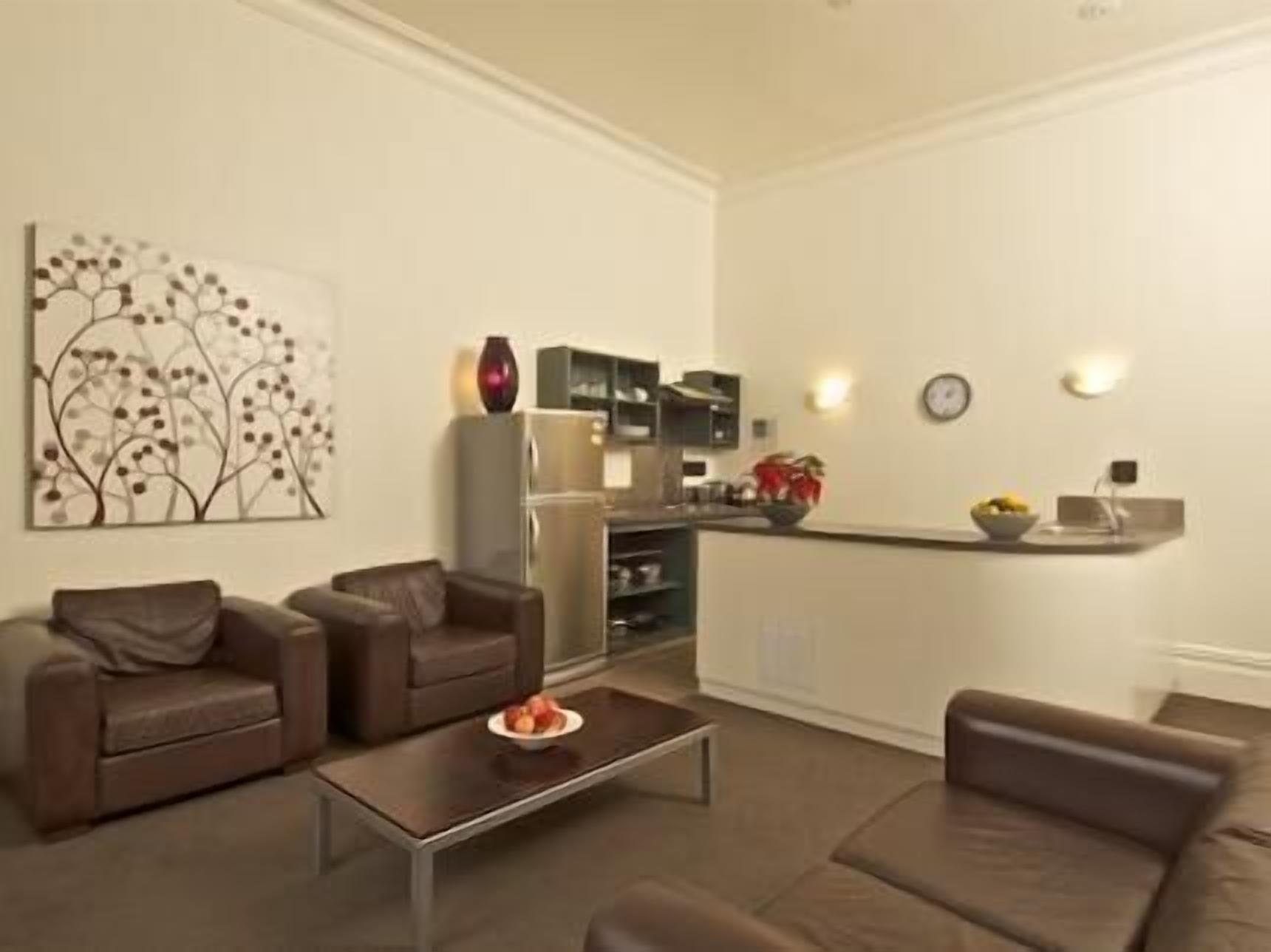 A modern living room with two brown sofas, a coffee table, and a kitchen area with a refrigerator, microwave, and a small island with a bowl of fruit.
