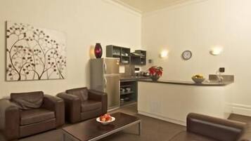 A modern living room with two brown sofas, a coffee table, and a kitchen area with a refrigerator, microwave, and a small island with a bowl of fruit.
