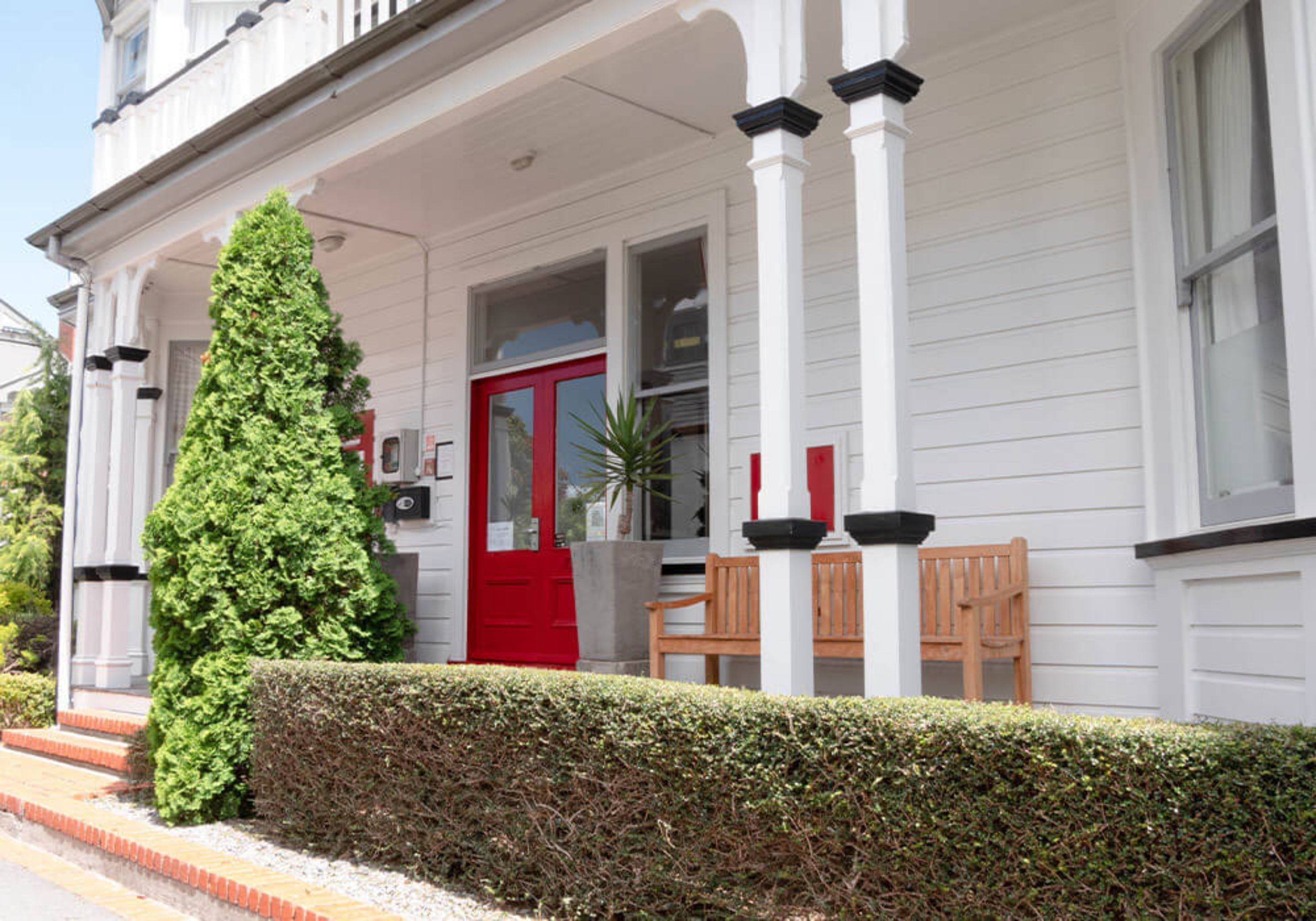 A white building with a red door, a wooden bench, and a tall green hedge.
