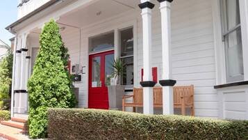 A white building with a red door, a wooden bench, and a tall green hedge.