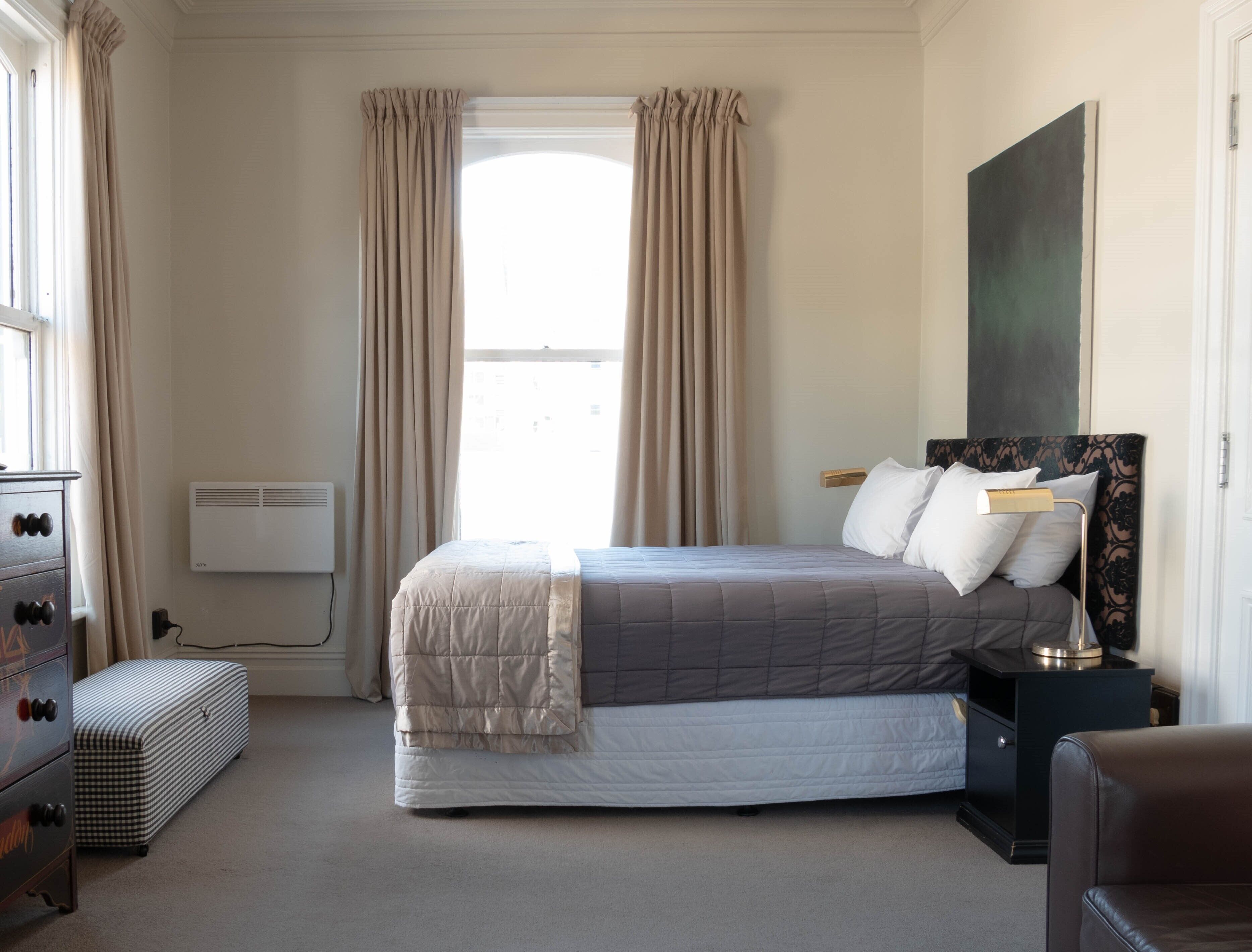 A bedroom with a bed, bedside table, dresser, and a large window with curtains.