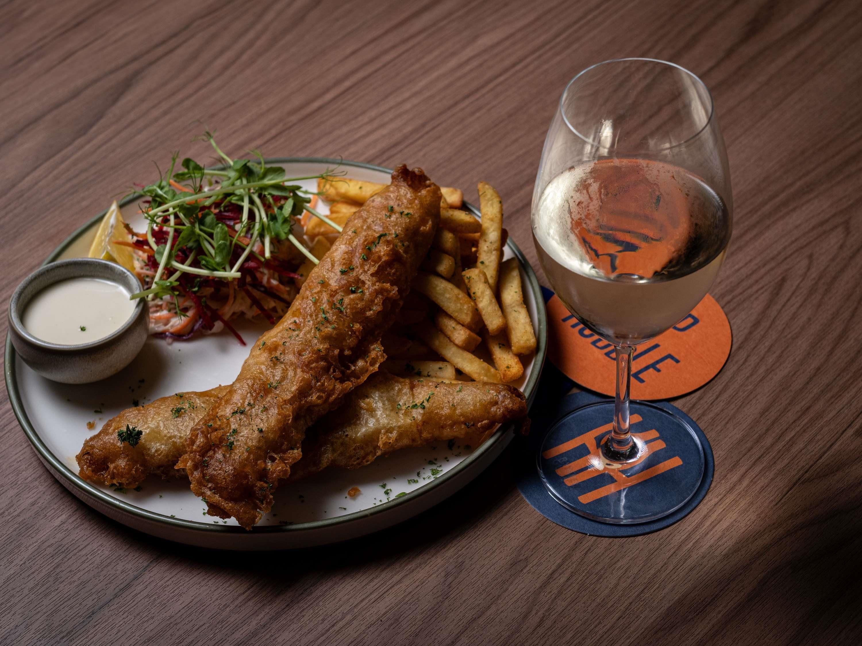 A plate of fish and chips with a side salad and a small bowl of sauce, accompanied by a glass of beer on a coaster.