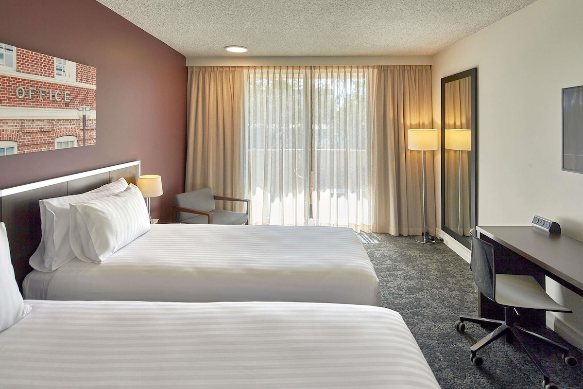 A hotel room with two beds, a desk, a chair, a lamp, and a large window with curtains.