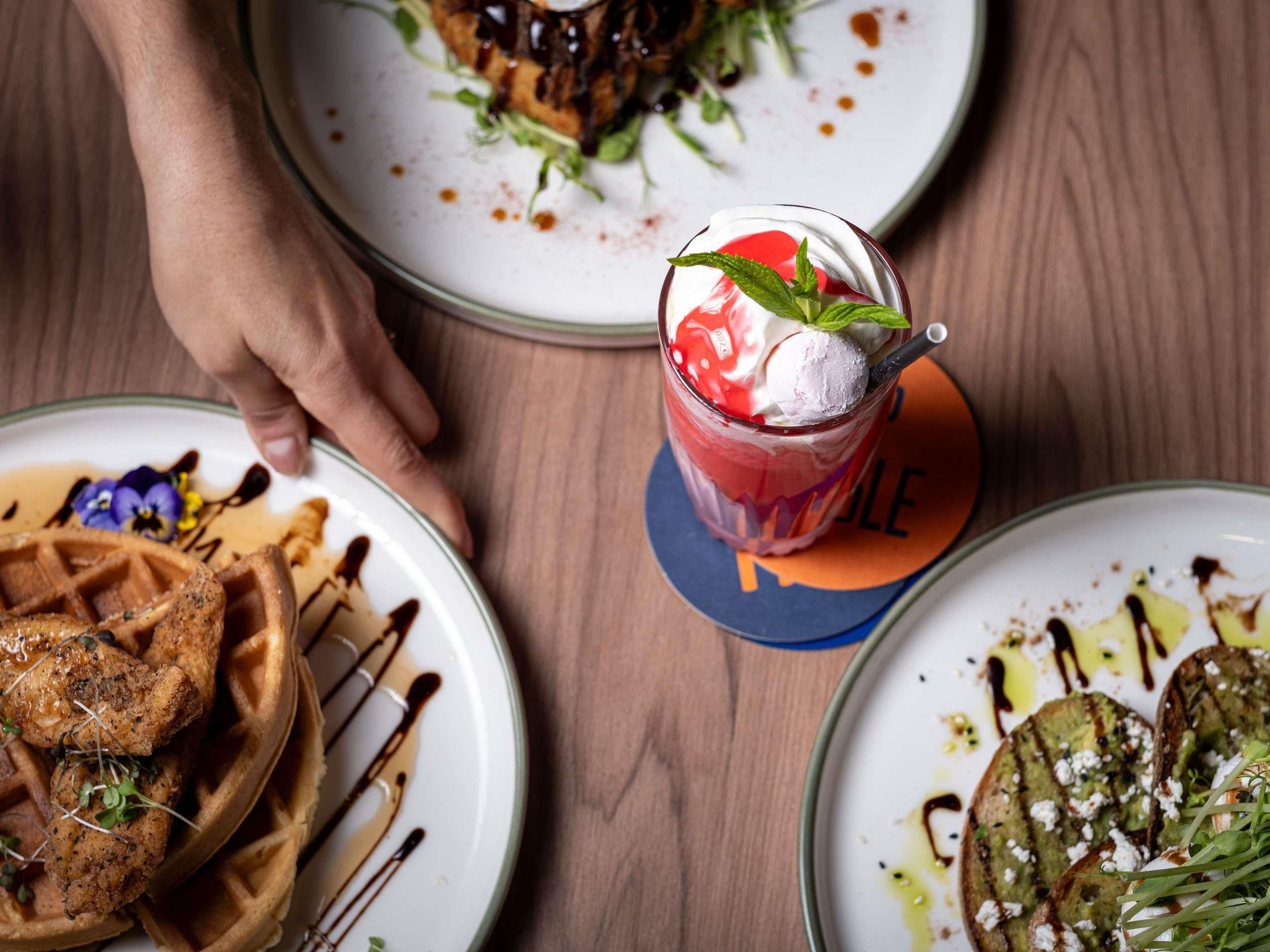 Une table avec des assiettes remplies de nourriture, notamment des gaufres et un verre d’une boisson rouge décorée d’une feuille de menthe.
