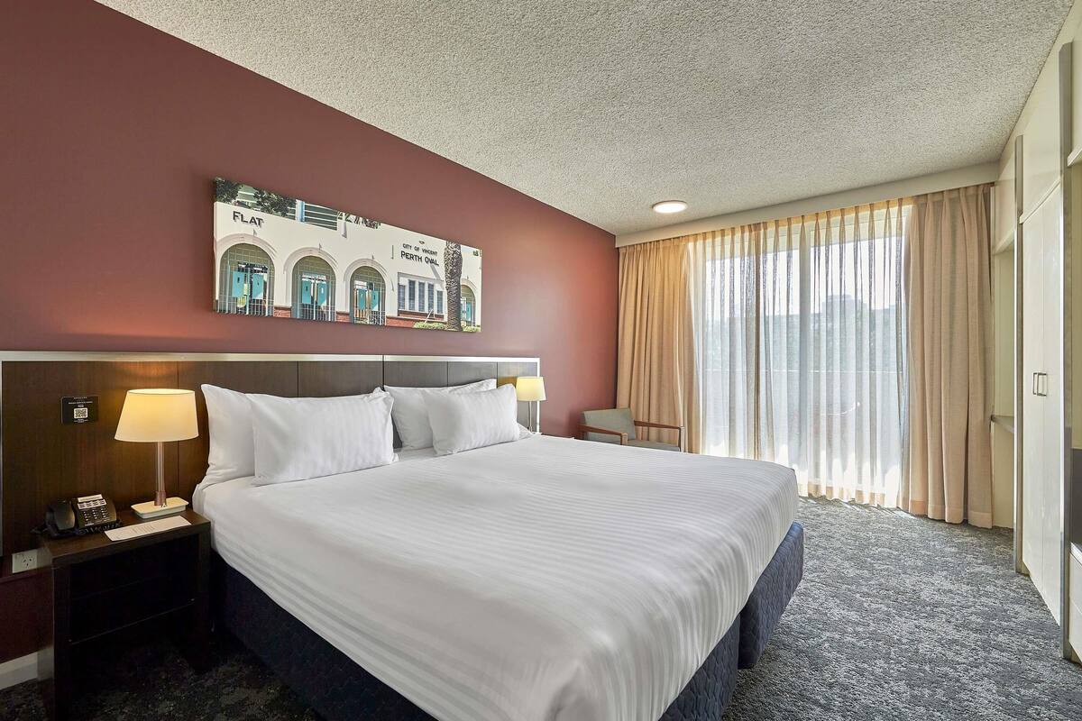 A hotel room with a large bed, a nightstand with a lamp, a chair, and a view of the cityscape through the window.
