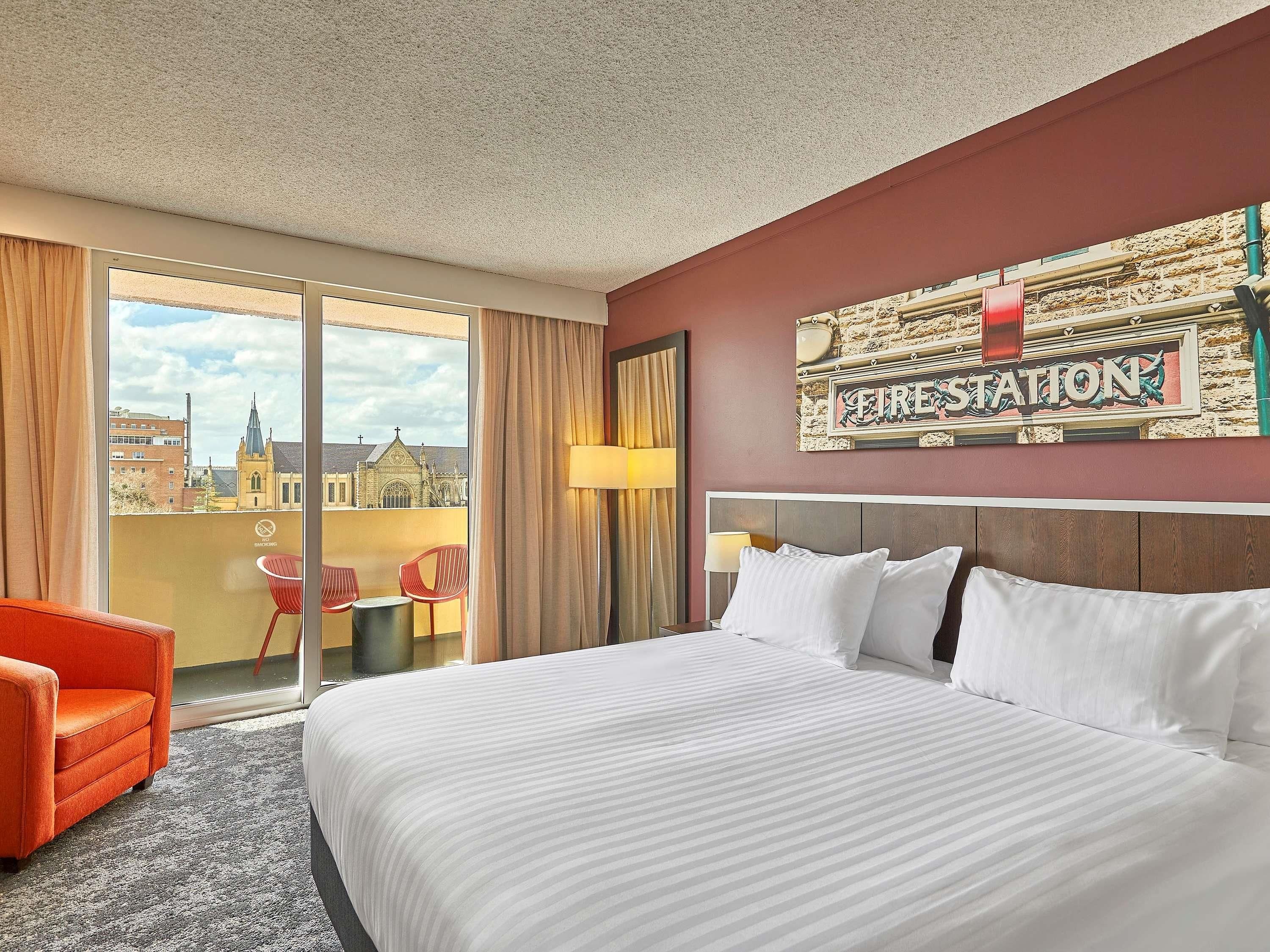 Quarto de hotel com uma cama grande, uma cadeira vermelha, vista para uma igreja e um aviso com a inscrição 'FIRE STATION'.