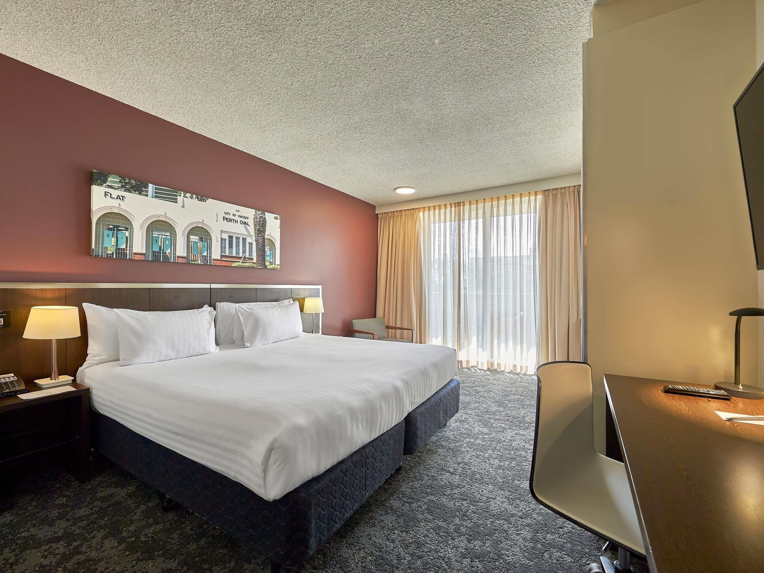 A hotel room with a bed, a desk, a chair, a lamp, a TV, and a painting on the wall.