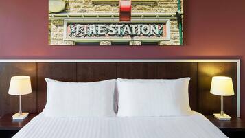 Une chambre d’hôtel avec un lit, deux lampes de chevet et une enseigne fixée au mur sur laquelle on peut lire « CASERNE DE POMPIERS ».