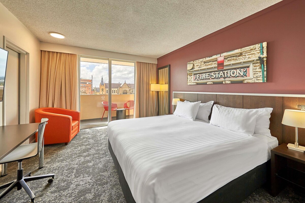 A hotel room with a large bed, a desk, a chair, and a view of a cityscape through the window.