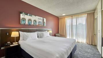 A hotel room with a large bed, a nightstand with a lamp, a chair, and a view of the cityscape through the window.