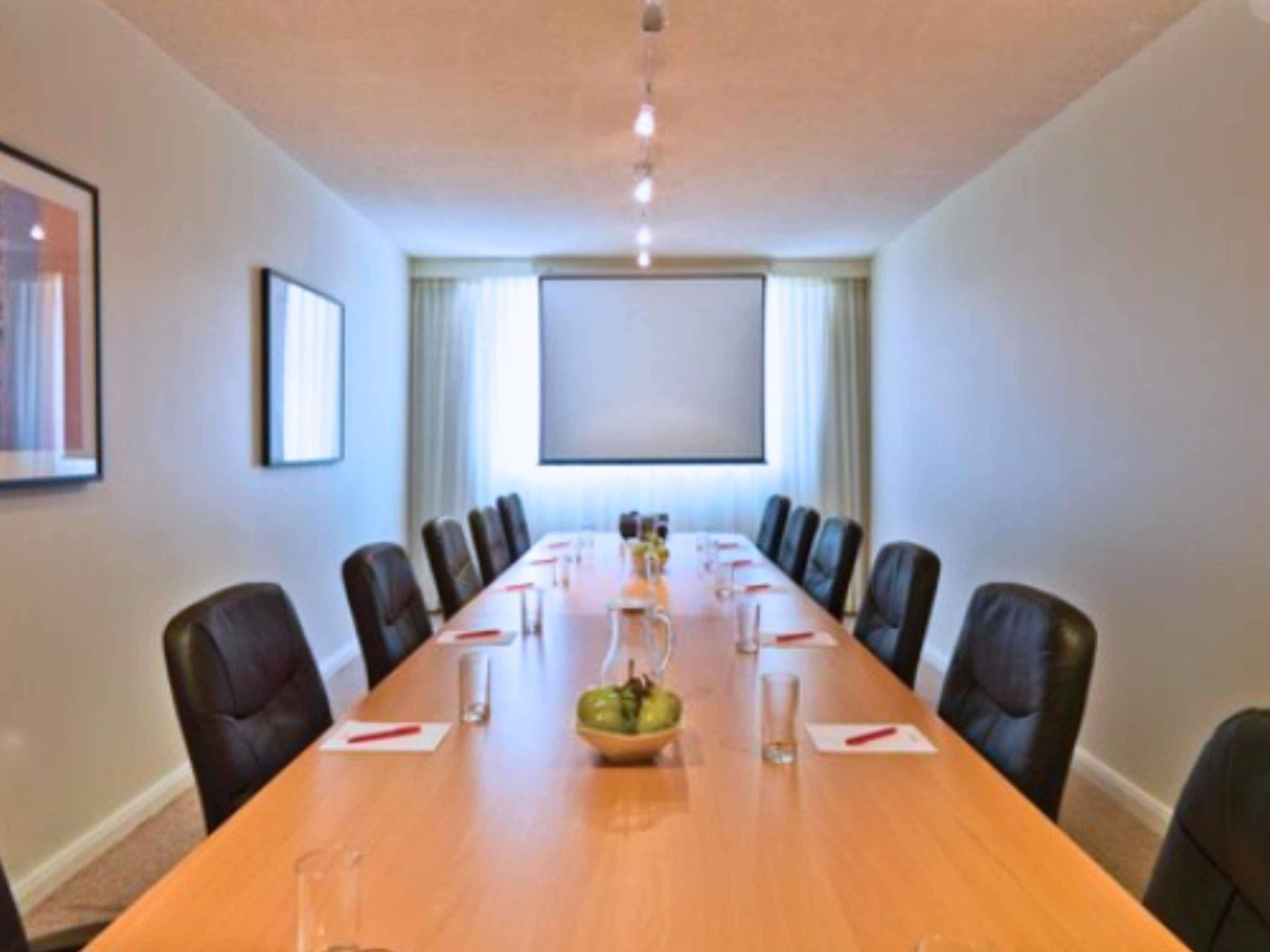 A conference room with a long table, chairs, a projector screen, and a window with curtains.