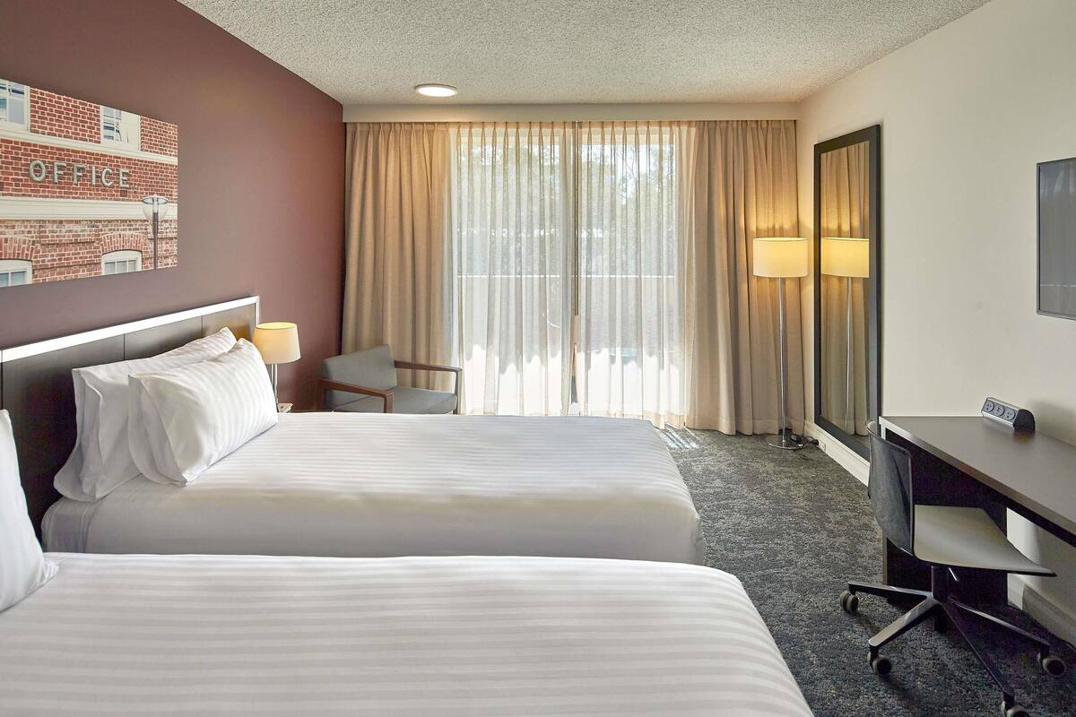 A hotel room with two beds, a desk, a chair, a lamp, and a large window with curtains.