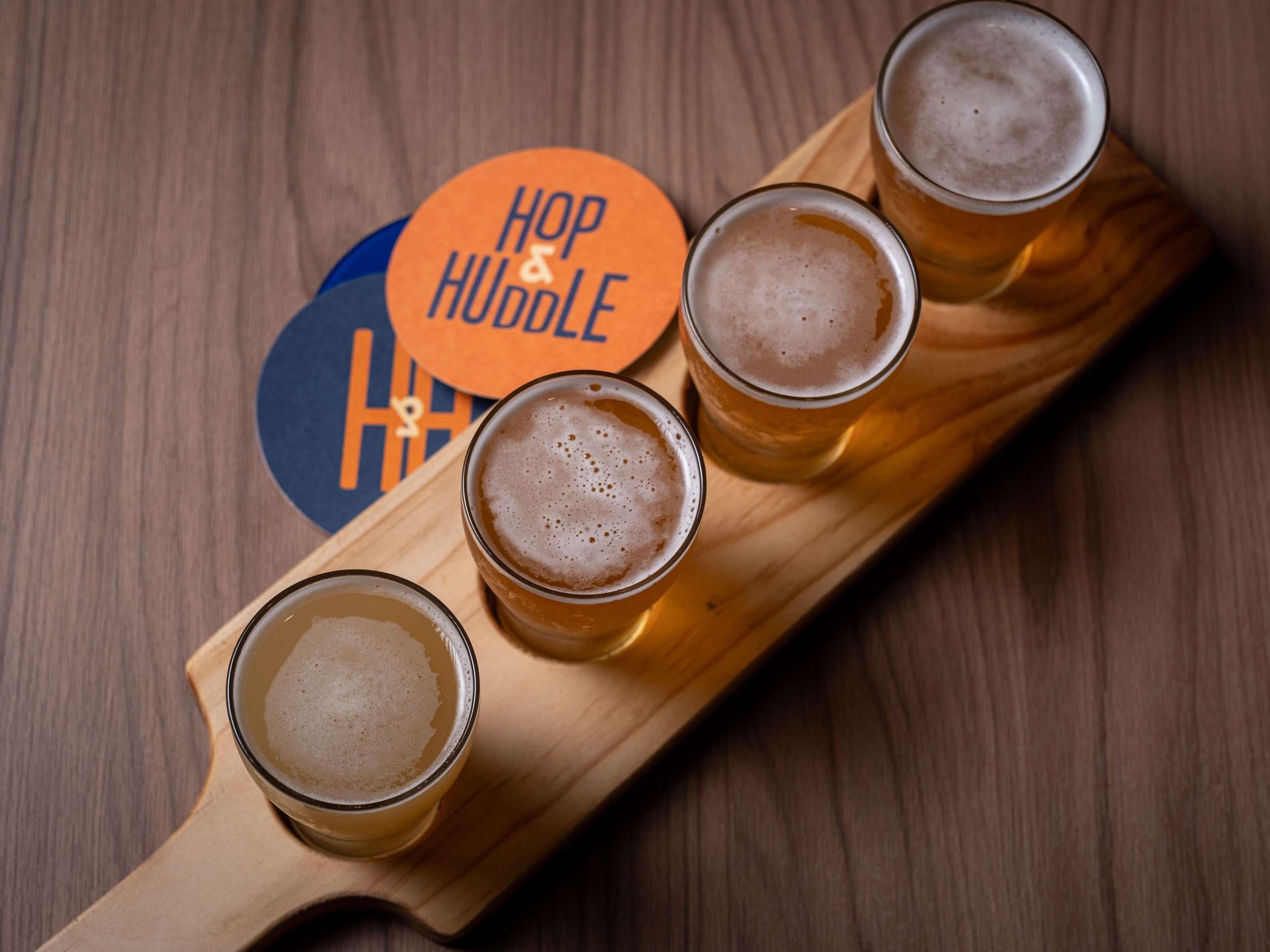 Four glasses of beer on a wooden tray with a coaster labeled 'Hop & Huddle.'