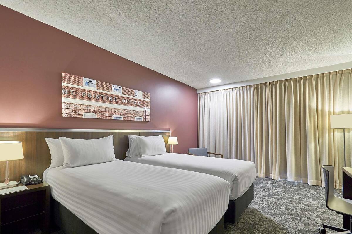 A hotel room with two beds, a desk, a chair, a lamp, and a painting on the wall.