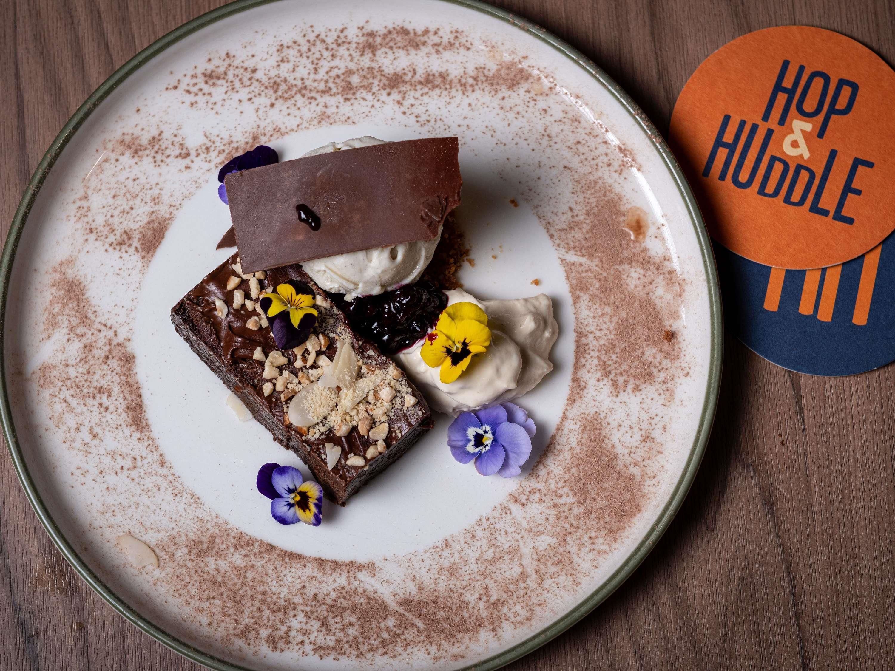 Une assiette à dessert avec un brownie, de la crème fouettée et un filet de chocolat, accompagnée d’un dessous de verre portant l’inscription « Hop & Huddle ».