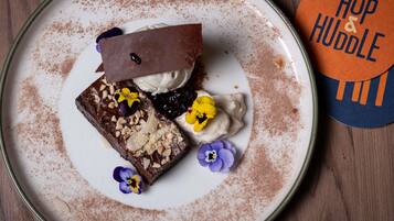 Une assiette à dessert avec un brownie, de la crème fouettée et un filet de chocolat, accompagnée d’un dessous de verre portant l’inscription « Hop & Huddle ».