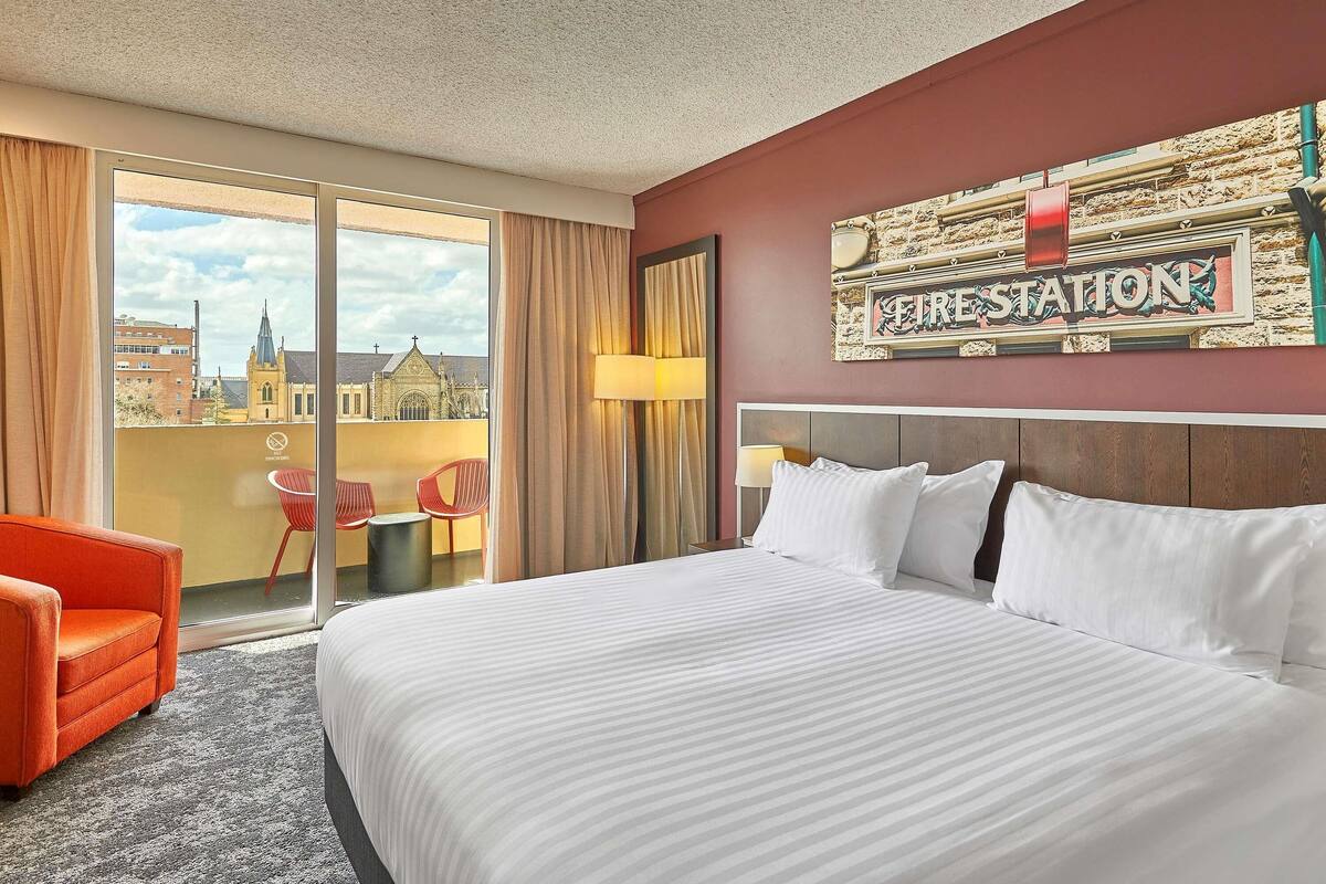 A hotel room with a large bed, a red chair, a view of a church, and a sign reading "FIRE STATION".