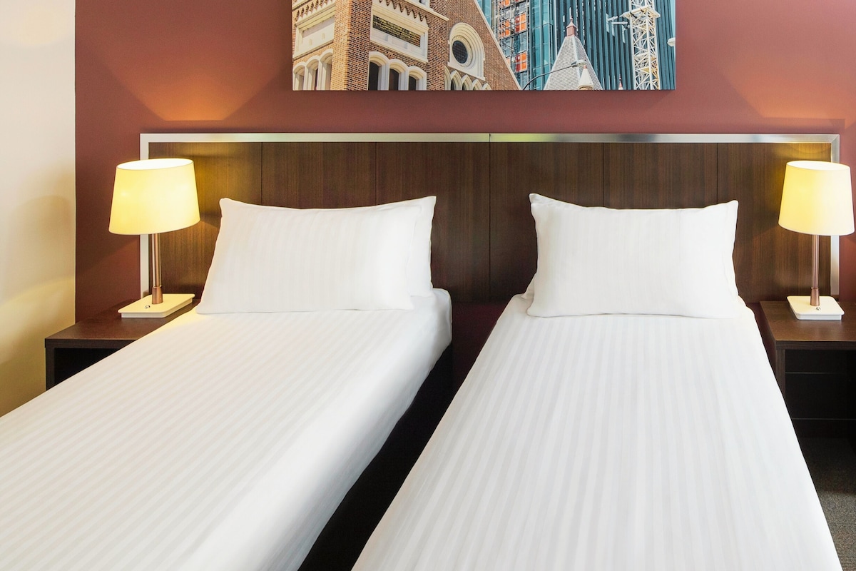 A hotel room with two single beds, each with a wooden headboard, white linens, and bedside lamps.