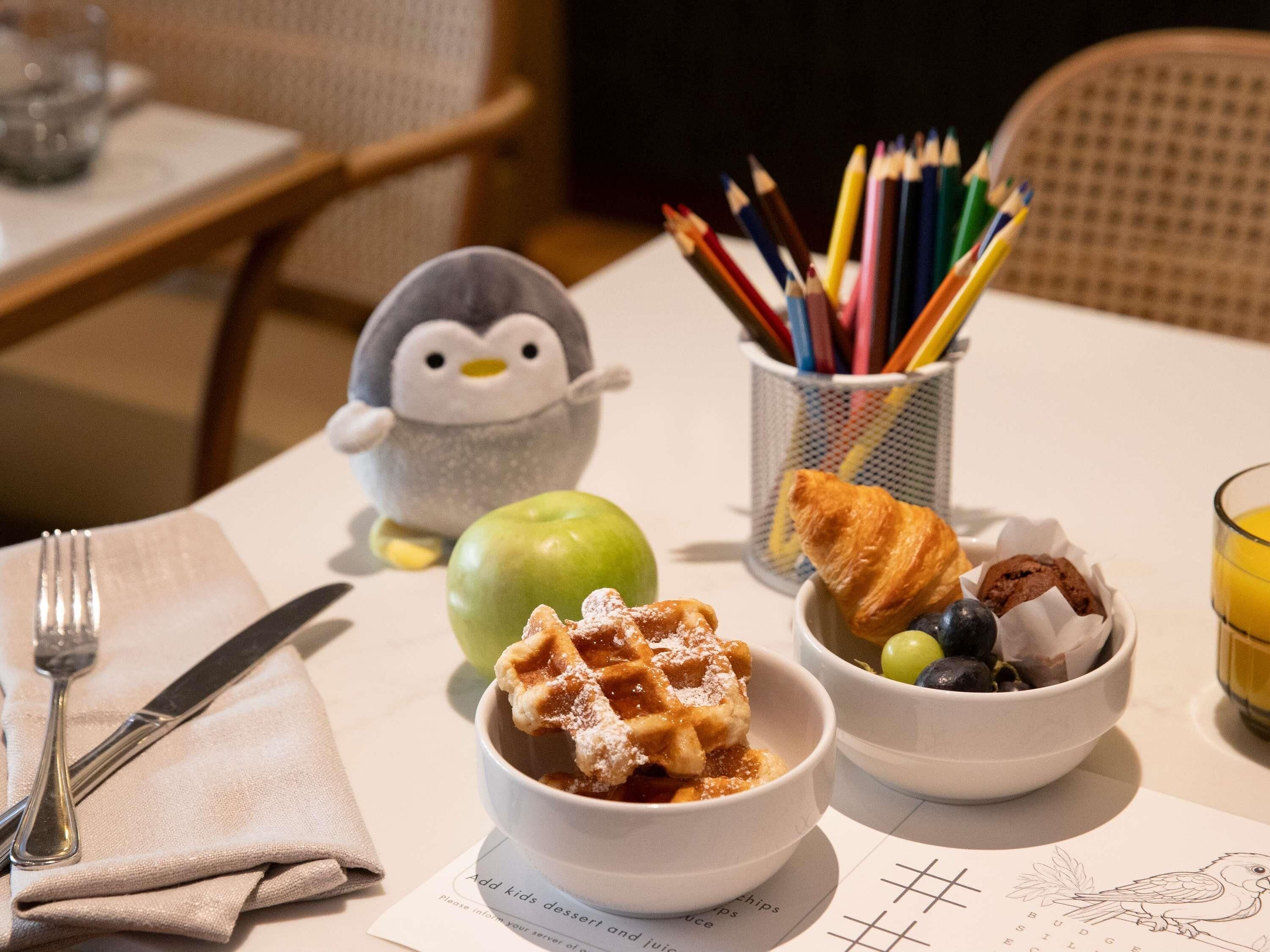 A breakfast setting with waffles, croissants, and fruit, a penguin toy, and a cup of colorful pencils.