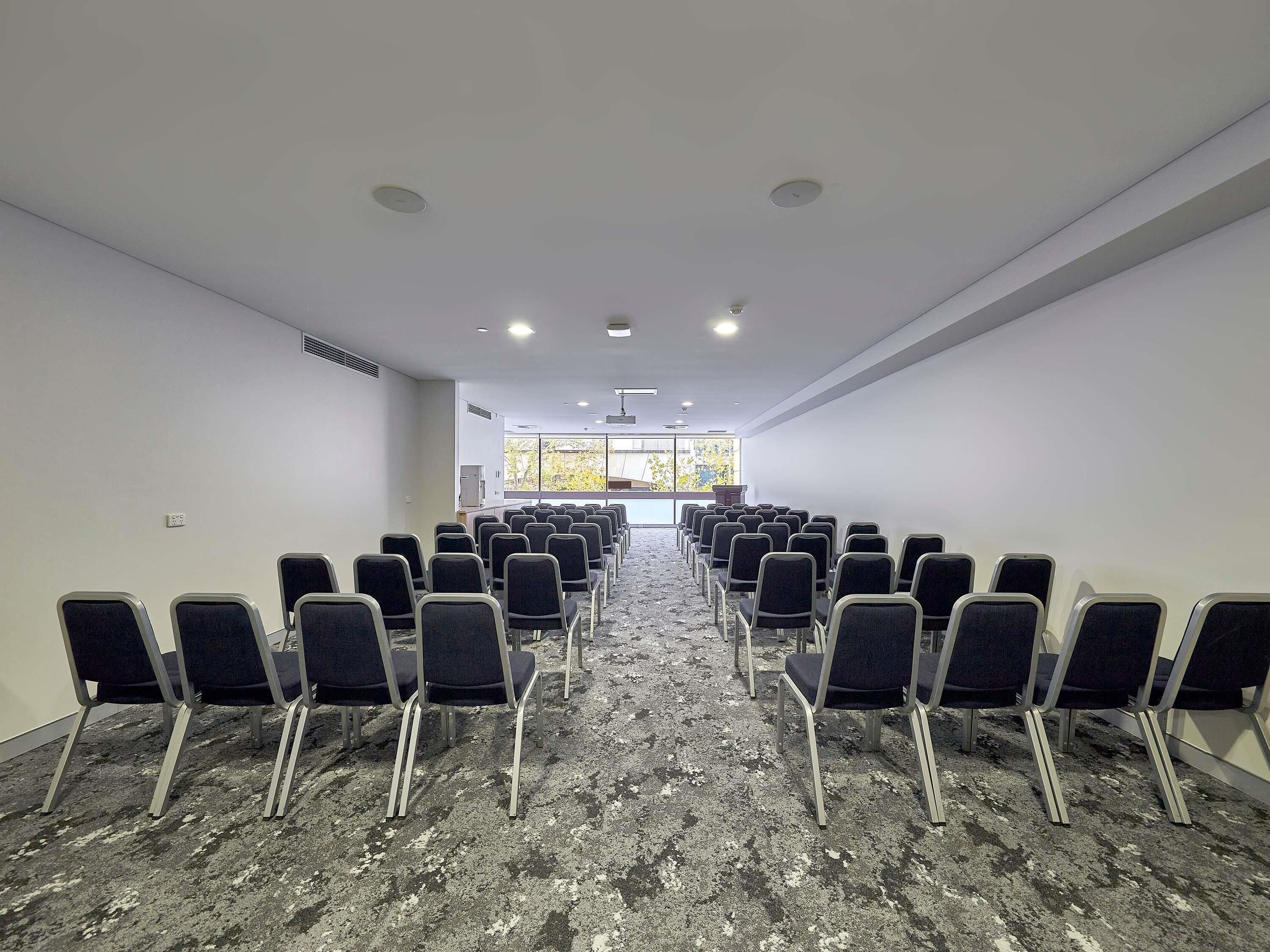 A conference room with rows of chairs facing a podium, large windows, and a carpeted floor.
