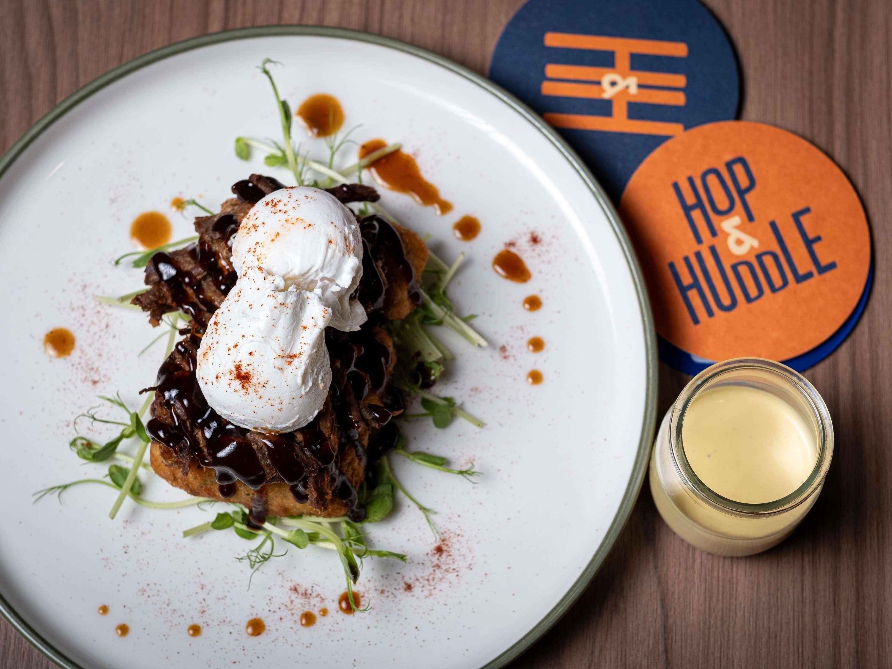 A plate of food with a poached egg, greens, and sauce, accompanied by a glass of a yellow beverage and a coaster with 'Hop & Huddle' text.