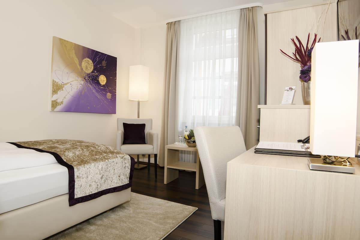Habitación de hotel con cama, escritorio, silla, lámpara, florero con flores y un cuadro en la pared.