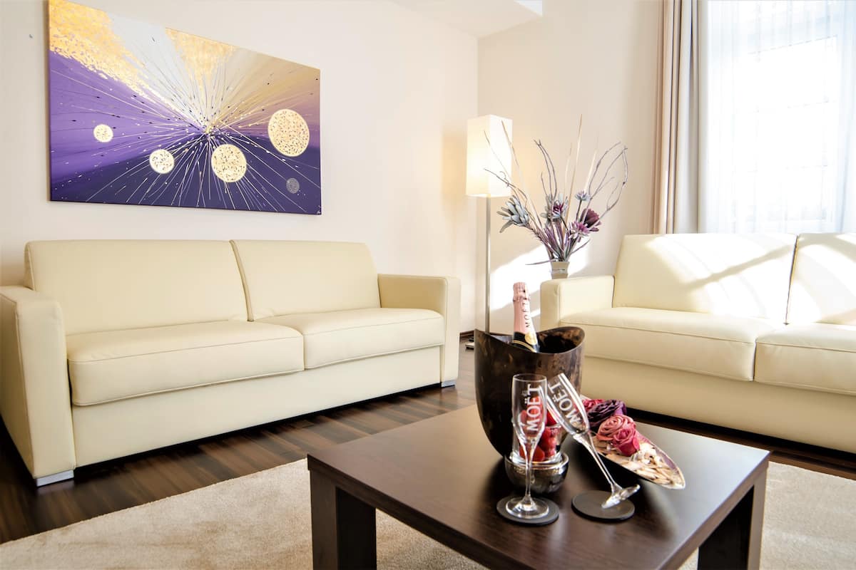 Un salón moderno con un sofá color beige, una mesa de centro de madera oscura con una botella de champán, una copa y un cuenco decorativo. Un cuadro abstracto grande adorna la pared, y una lámpara de pie está junto al sofá.