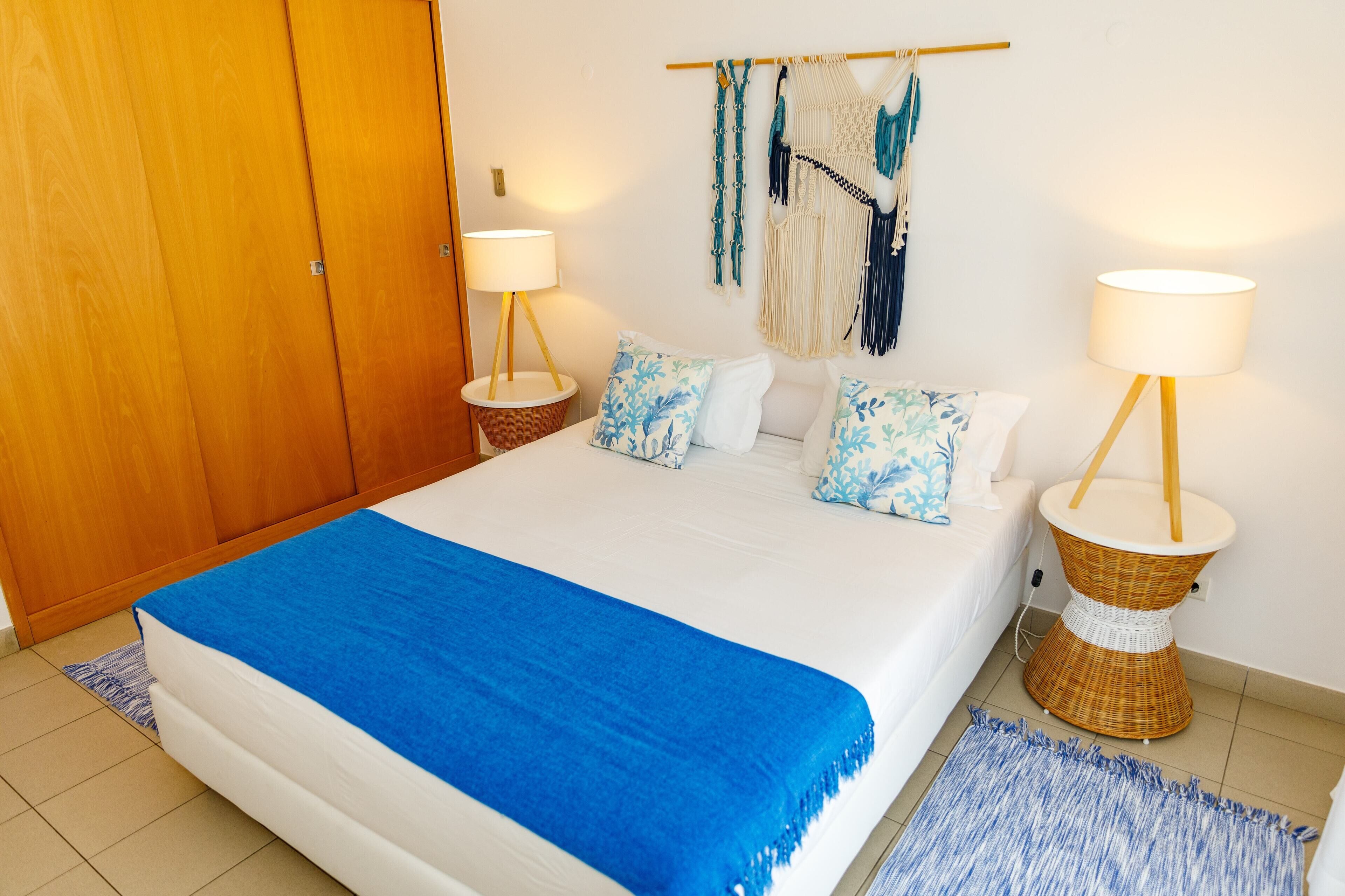 A neatly made bed with a blue blanket, two bedside lamps, and a macrame wall hanging.