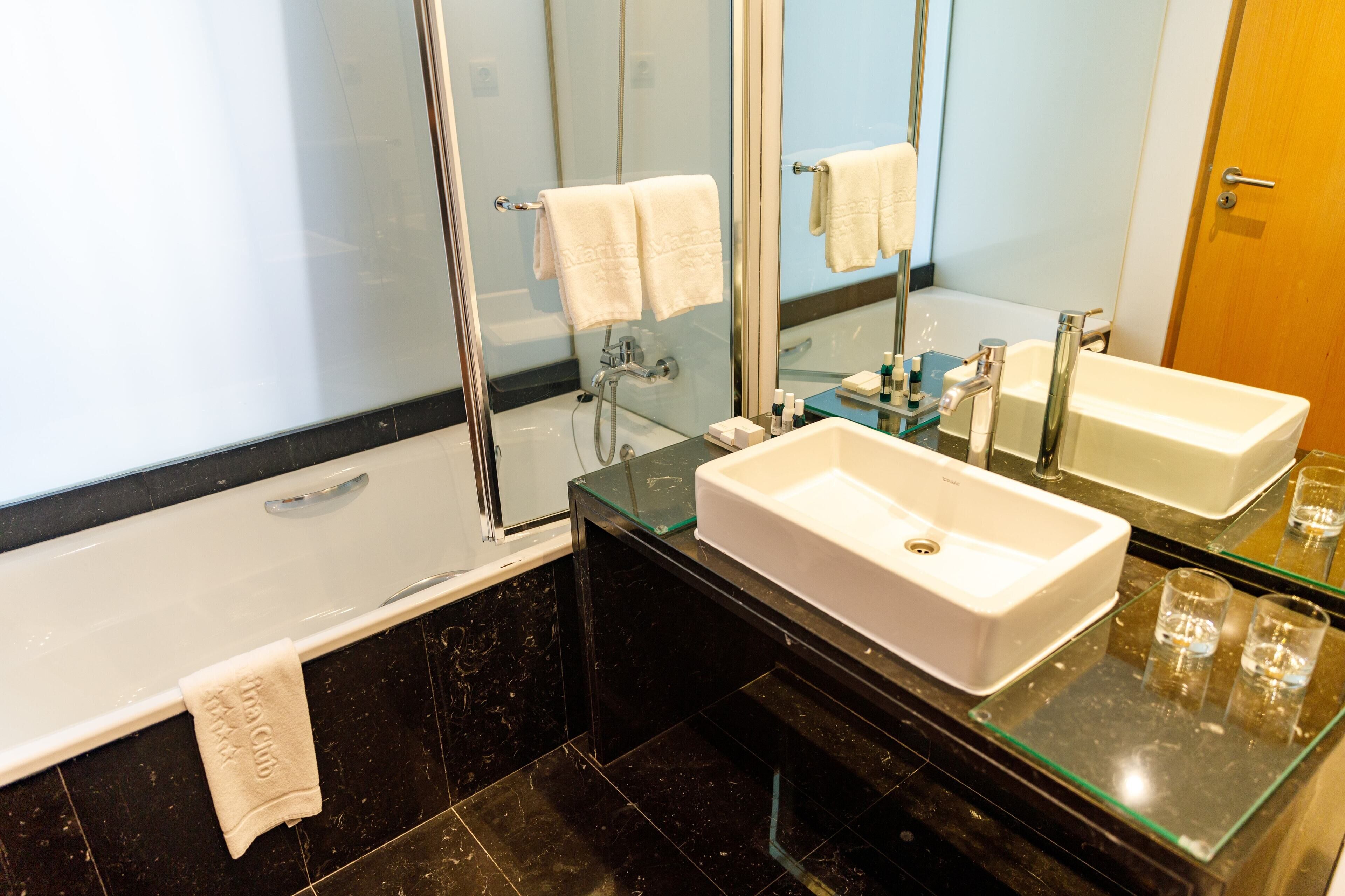 A modern bathroom with a glass-enclosed shower, a white bathtub, and a glass countertop with a white sink.