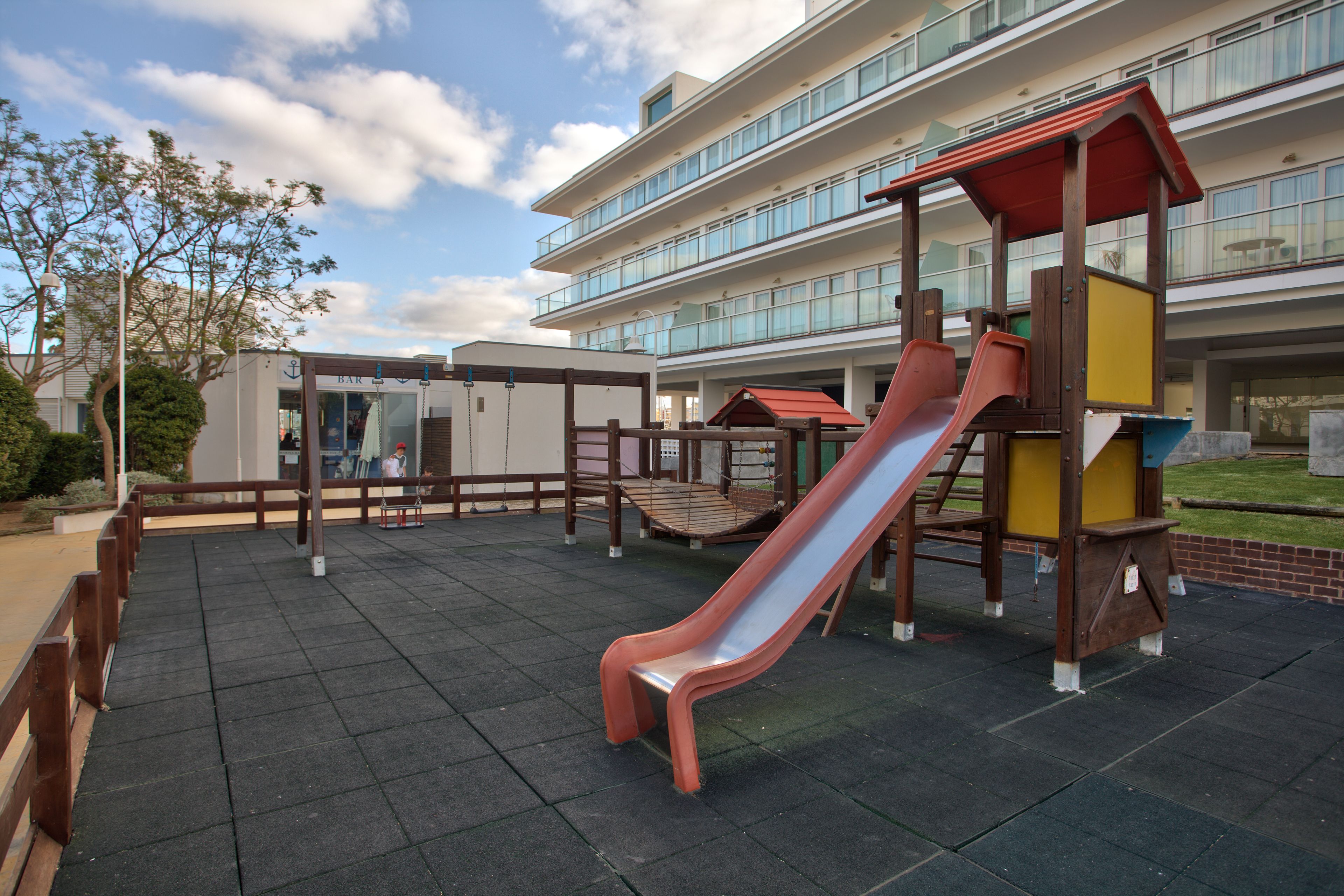 A playground with a slide and climbing structures, surrounded by a building with multiple floors and large windows.