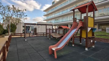 A playground with a slide and climbing structures, surrounded by a building with multiple floors and large windows.