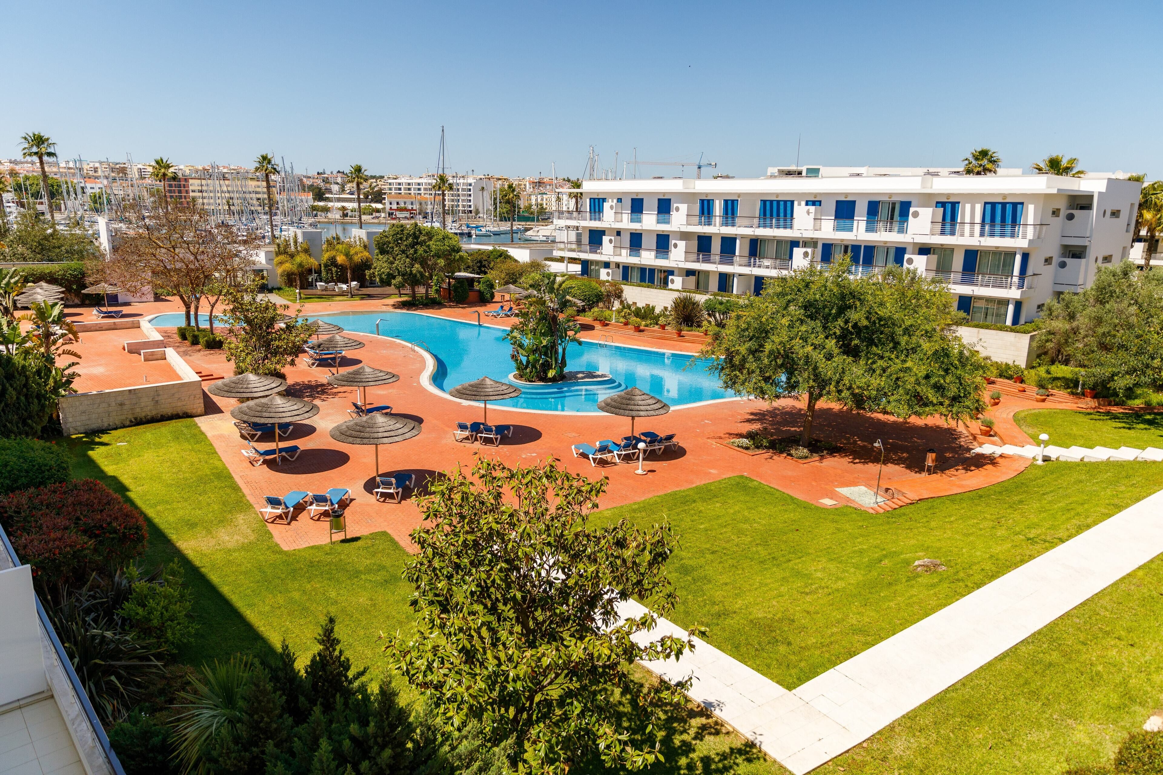 A hotel with a swimming pool, sun loungers, and a view of the marina.