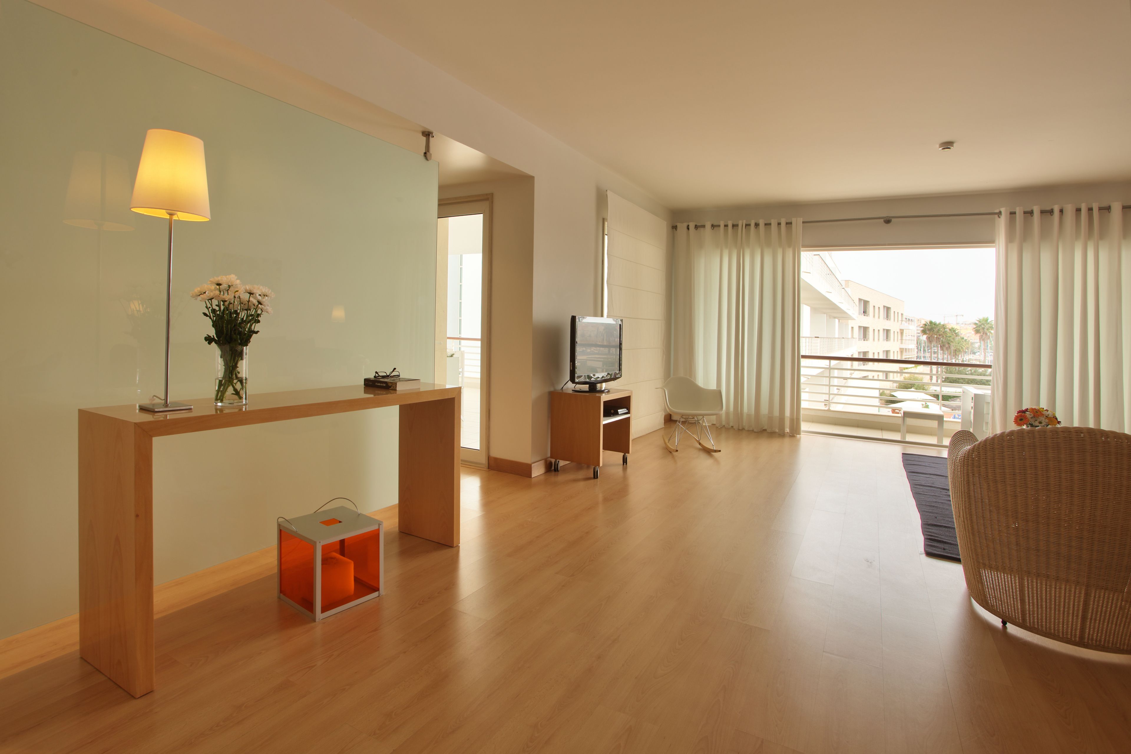 Ein modernes Hotelzimmer mit einem Holztisch, einem Fernseher, einem Korbstuhl und einem Balkon mit Blick auf Gebäude und Palmen.