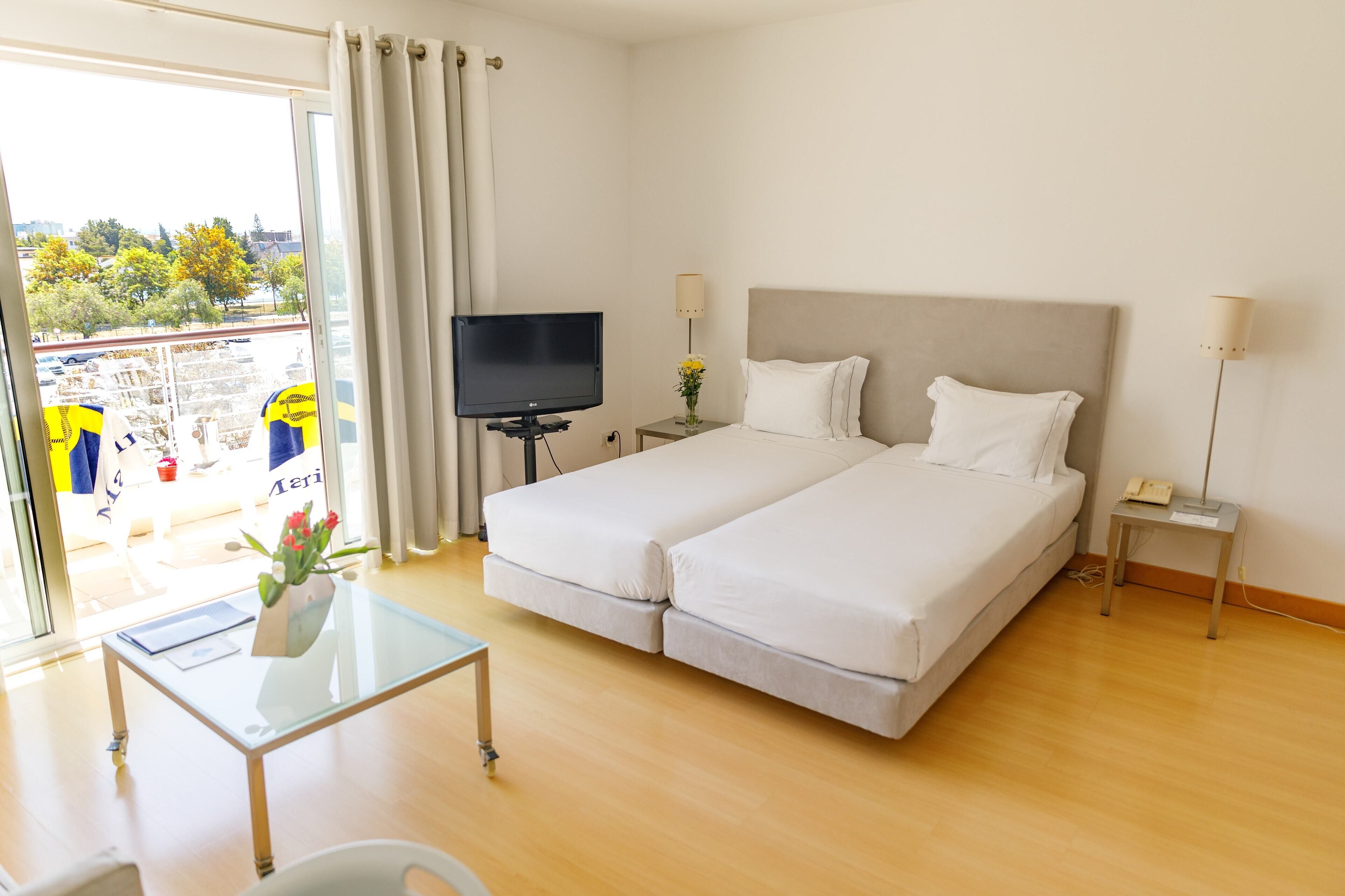 A hotel room with a bed, a TV, a bedside table, a lamp, and a view of the outside.