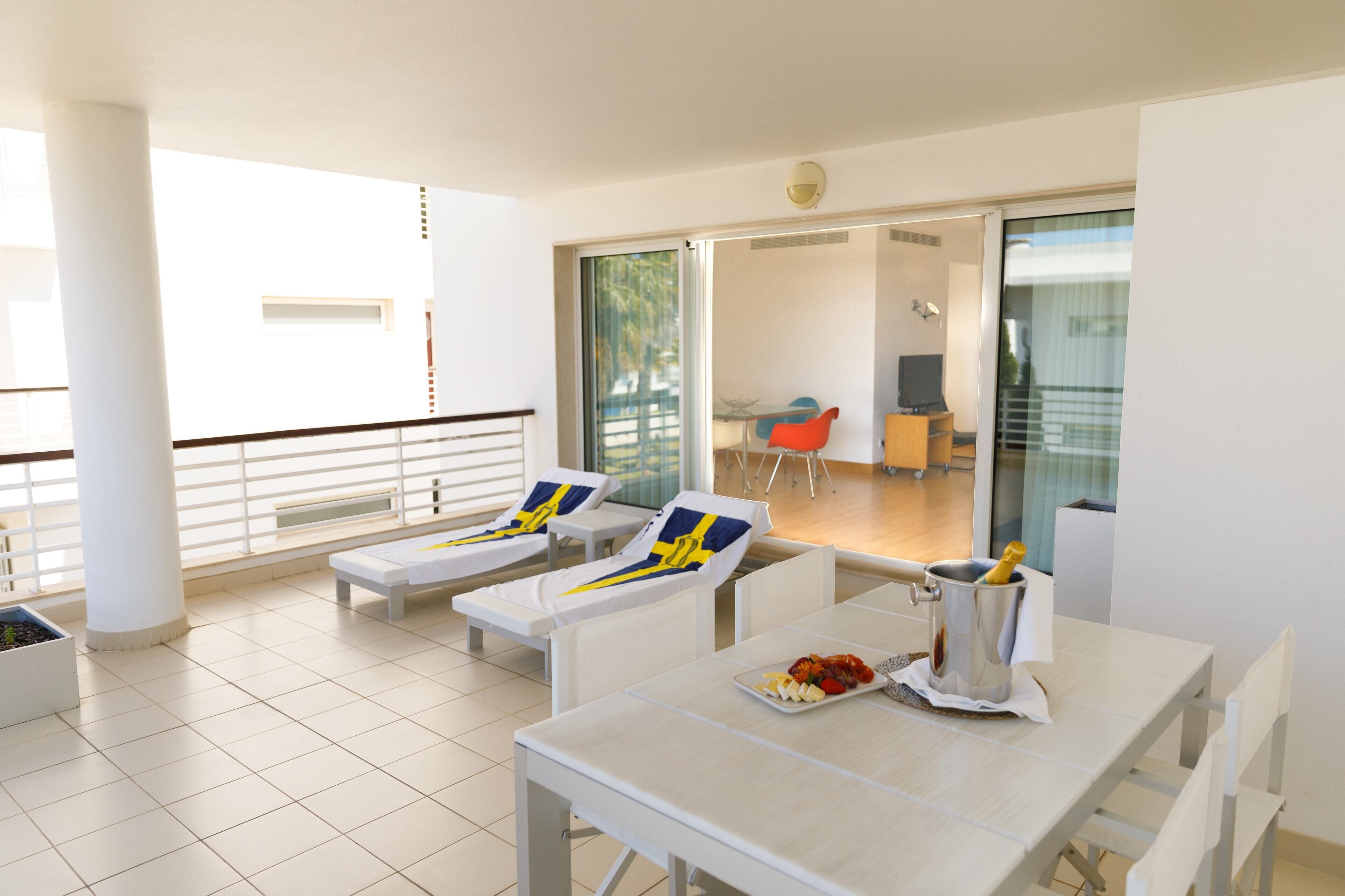 Una habitación de hotel moderna con balcón, muebles blancos y zona de comedor con mesa dispuesta para dos personas.
