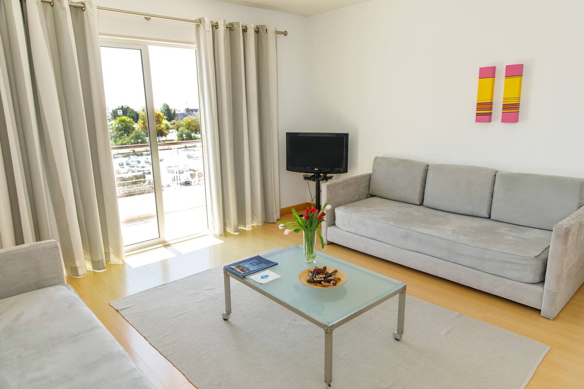 Ein modernes Wohnzimmer mit einem grauen Sofa, einem Glastisch mit einem Buch und einer Schüssel Essen, einem Fernseher auf einem Schrank und einem Balkon mit Vorhängen.