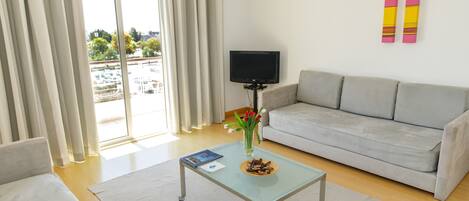 A modern living room with a grey sofa, a glass coffee table with a book and a plate of food, a TV on a stand, and a balcony with curtains.