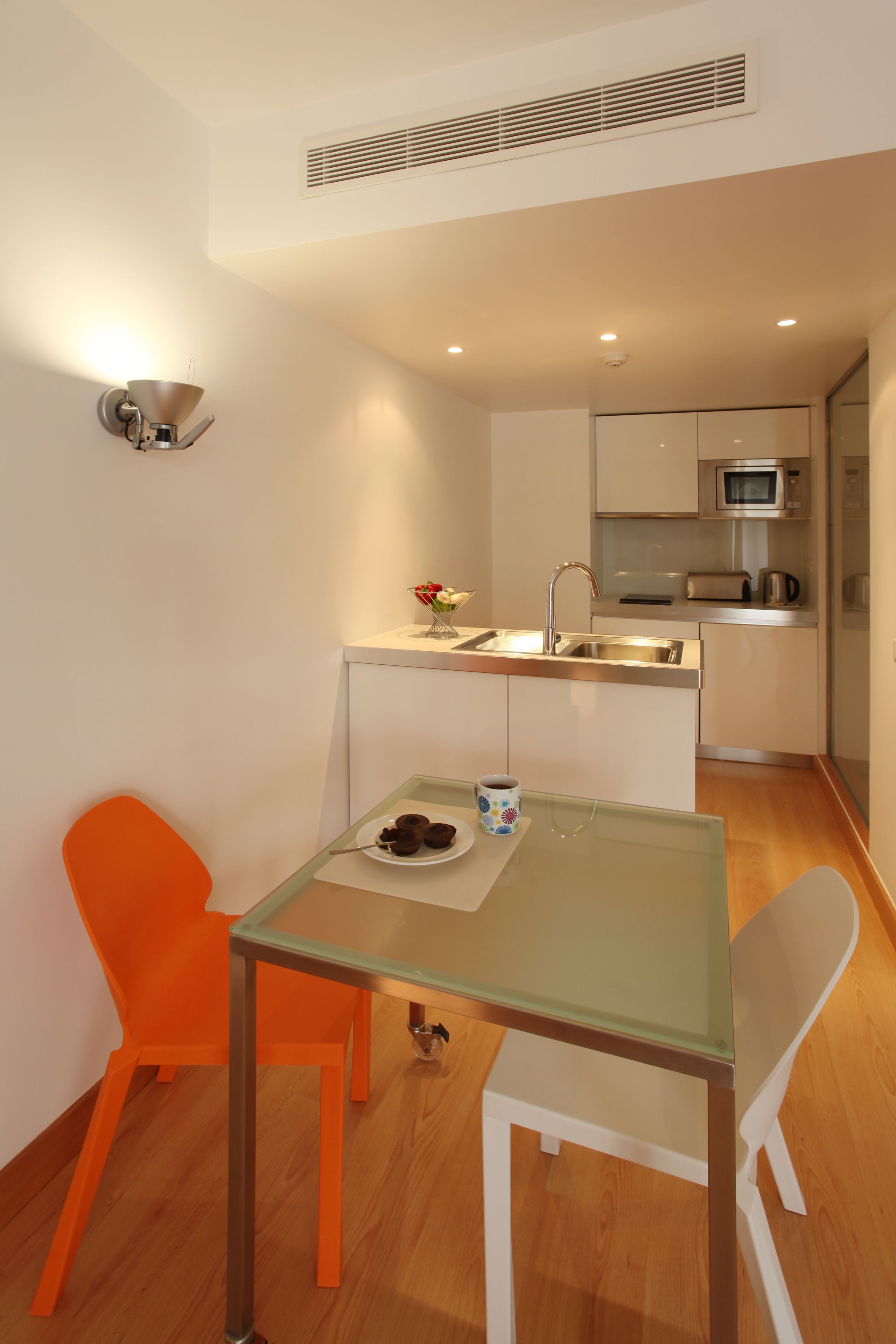 A compact kitchen with a microwave, toaster, and sink. There is a small dining area with a glass table and two chairs, one orange and one white.