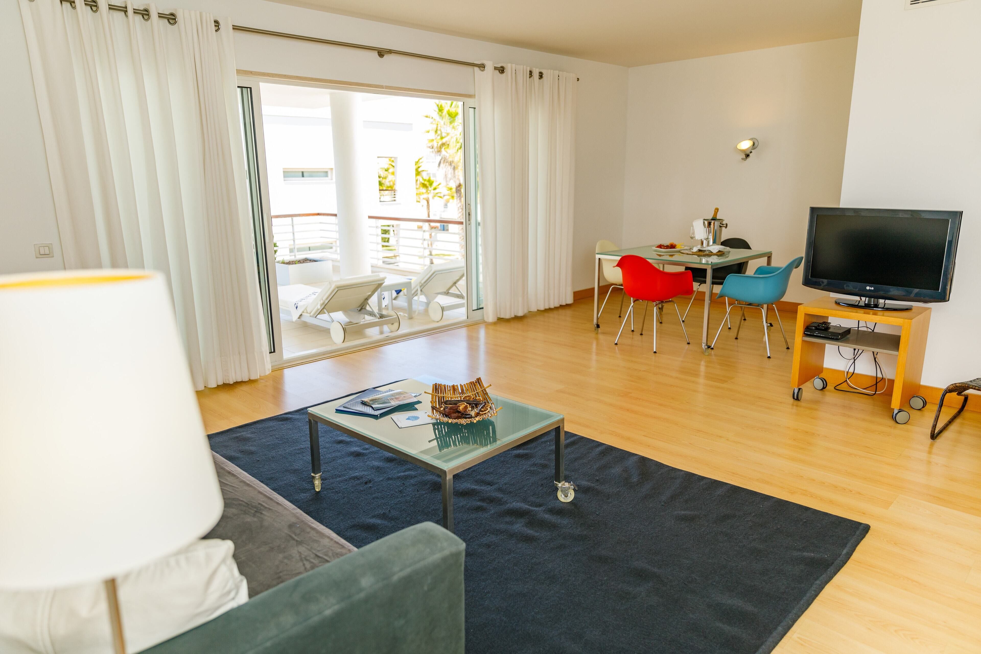 A modern living room with a sofa, a coffee table, a TV on a wooden stand, and a dining area with chairs and a table.