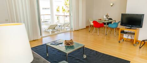 A modern living room with a sofa, a coffee table, a TV on a wooden stand, and a dining area with chairs and a table.