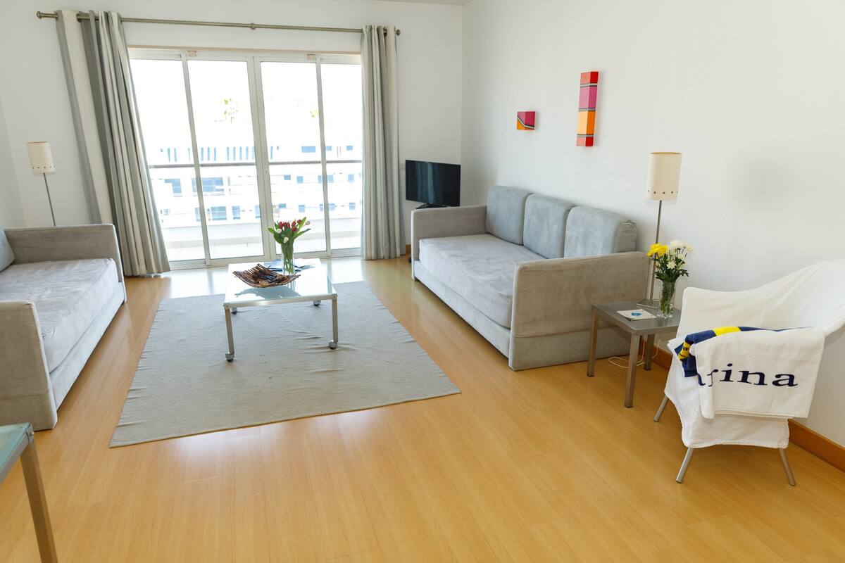 Ein modernes Hotelzimmer mit einer Couch, einem kleinen Tisch mit Blumenvase, einem Sessel mit Handtuch und einem Fernseher auf einem Schrank.