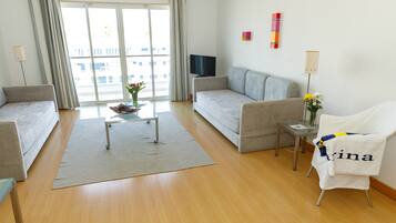A modern hotel room with a sofa, a small table with a vase of flowers, a chair with a towel, and a TV on a stand.