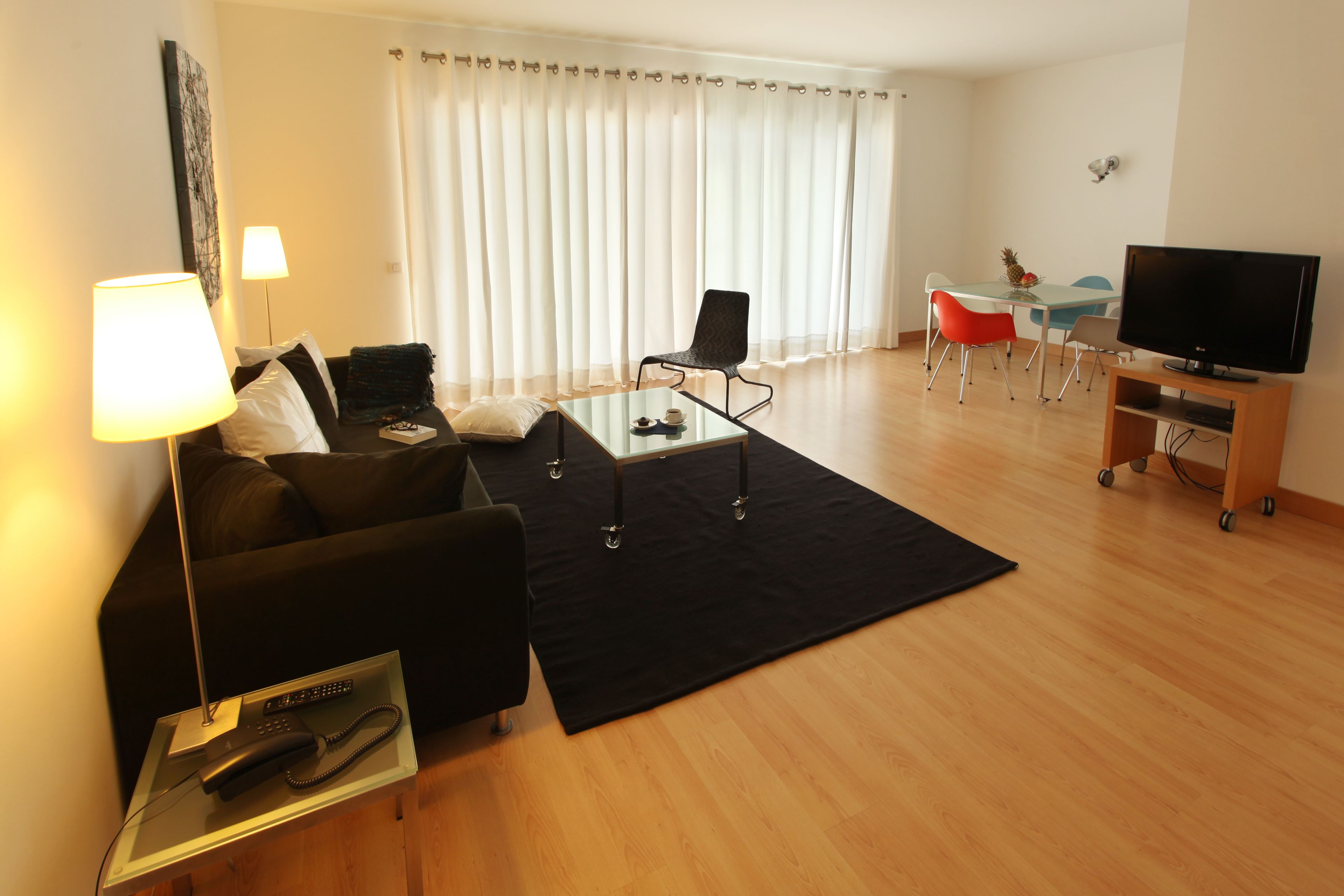 A modern living room with a black sofa, a glass coffee table, a TV on a wooden stand, and a dining area with a table and chairs.